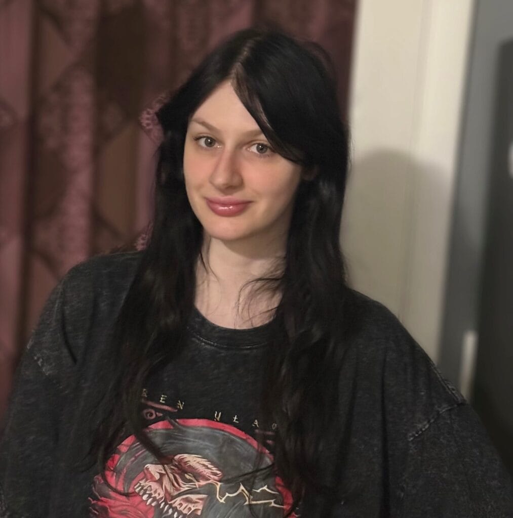 A young woman with long, dark hair stands indoors, smiling softly. She is wearing a loose, dark graphic T-shirt with a dragon design. The background features patterned curtains and a white doorframe.