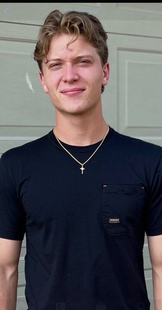 A young man with light brown hair wearing a black t-shirt and a gold cross necklace stands in front of a light green garage door, smiling slightly at the camera.