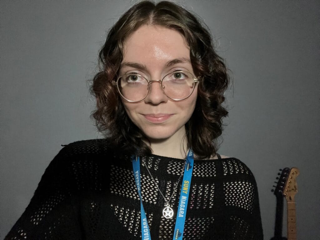 A person with wavy brown hair and glasses smiles at the camera. They wear a black knit top, a necklace, and a blue lanyard that says SUNY Niagara. A guitar is partially visible in the background.