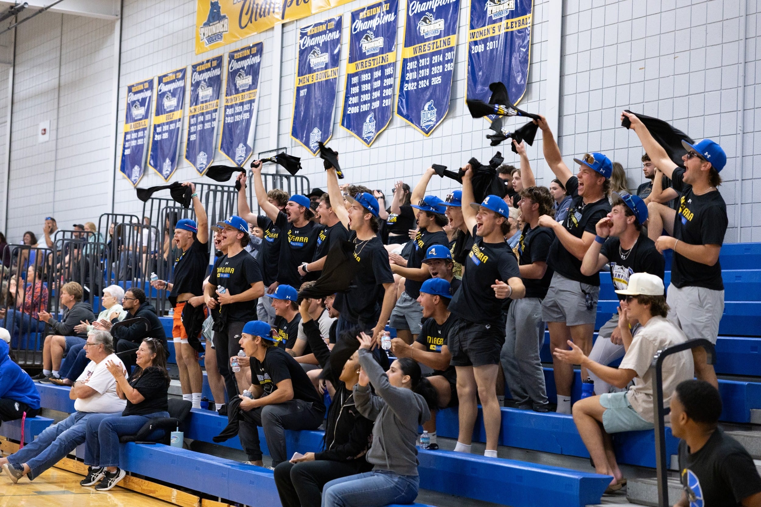 A group of enthusiastic fans wearing matching shirts and blue hats cheer and wave towels in the bleachers of a gymnasium during a sporting event. Banners hang on the wall above them.