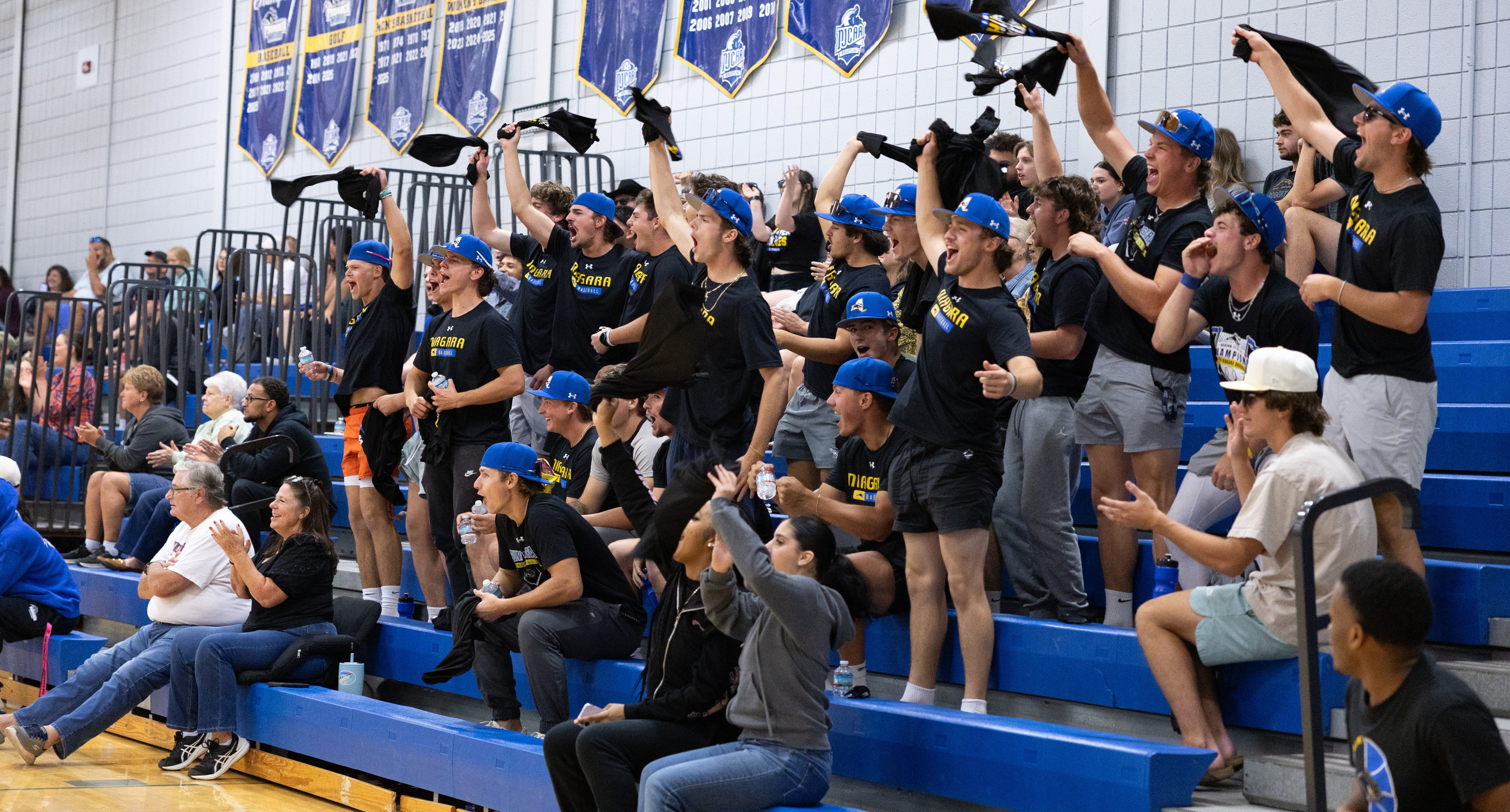A group of enthusiastic fans in matching shirts and blue hats cheer and wave shirts in the stands of an indoor gymnasium, while other spectators sit and watch. Sports banners hang on the wall above them.