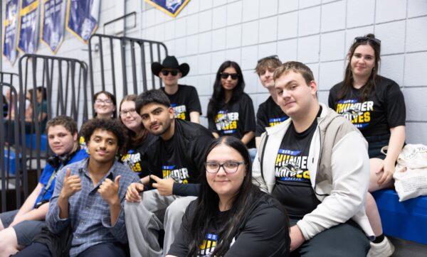 A group of students sit together on bleachers in a gym, many wearing matching black Thunder Wolves shirts. Some smile and give thumbs up, while others sit relaxed, posing for the photo.