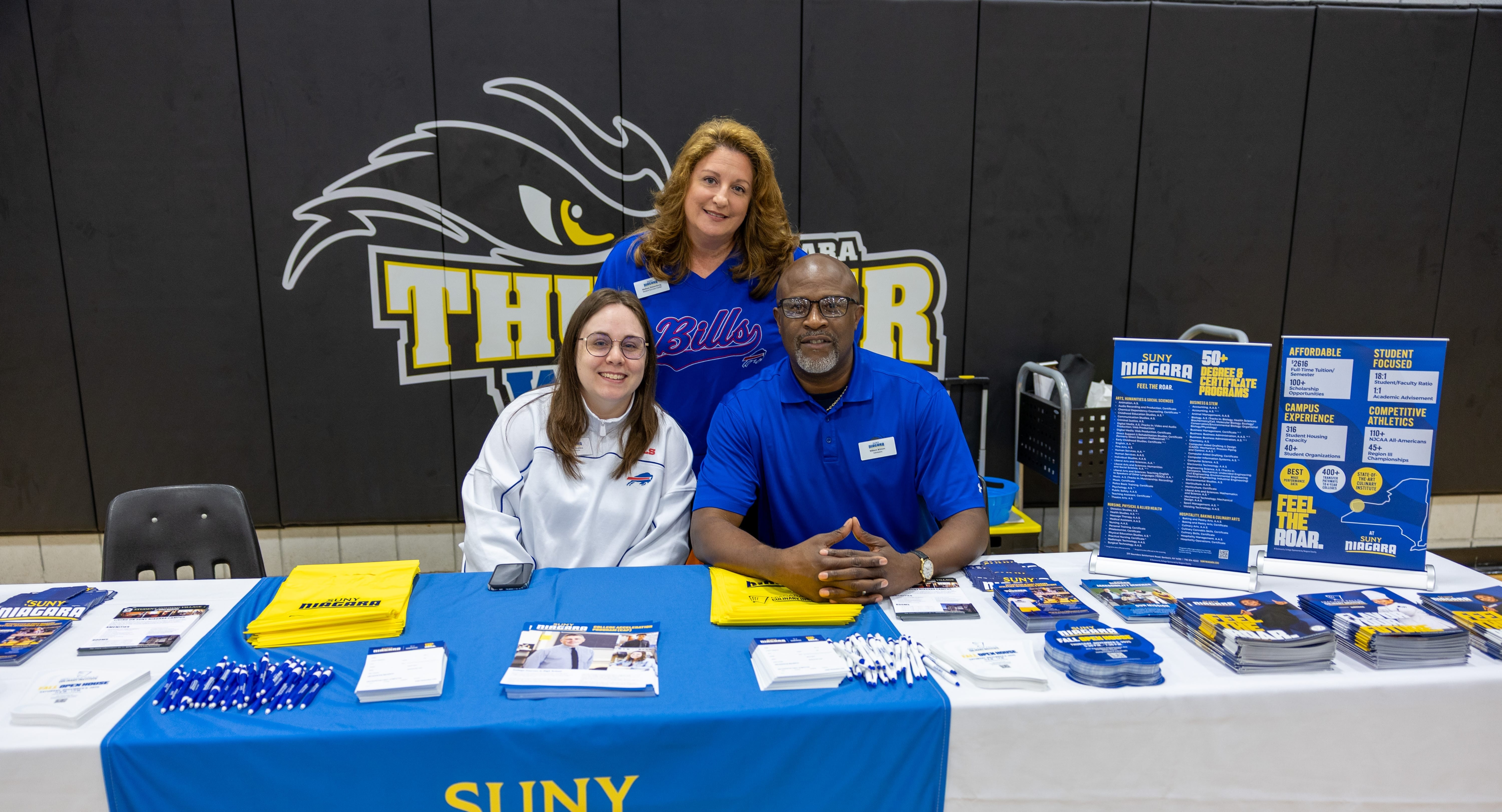 Three people smile at a SUNY information booth covered with pamphlets, flyers, pens, and other materials. A Thunder Wolves logo is on the wall behind them in a gymnasium setting.