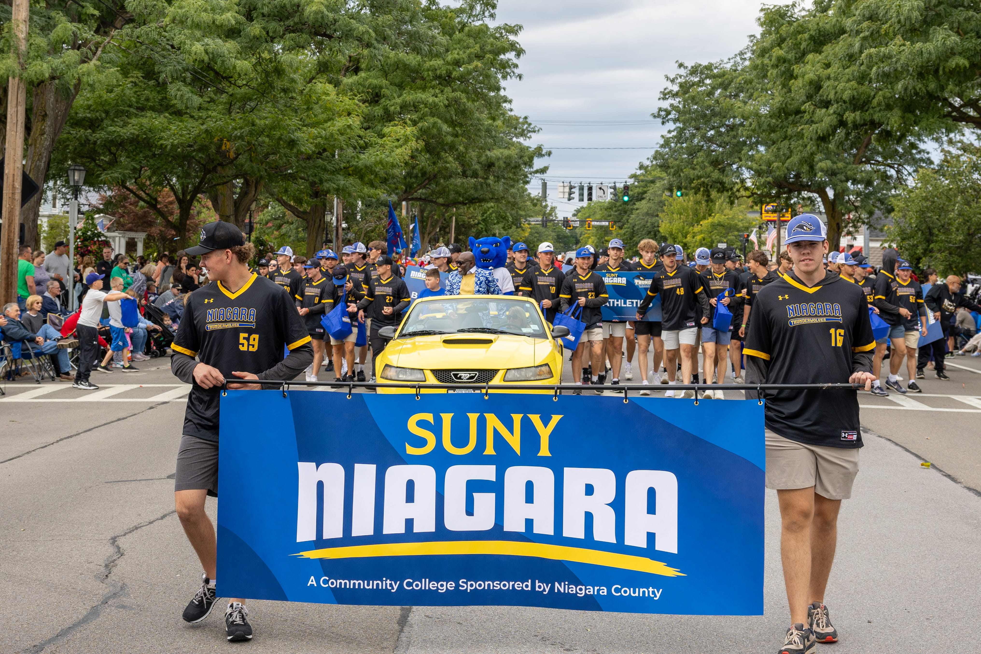 A group of people in matching sports jerseys marches in a parade, holding a blue SUNY Niagara banner. A yellow car and more participants follow behind on a tree-lined street, with spectators on both sides.