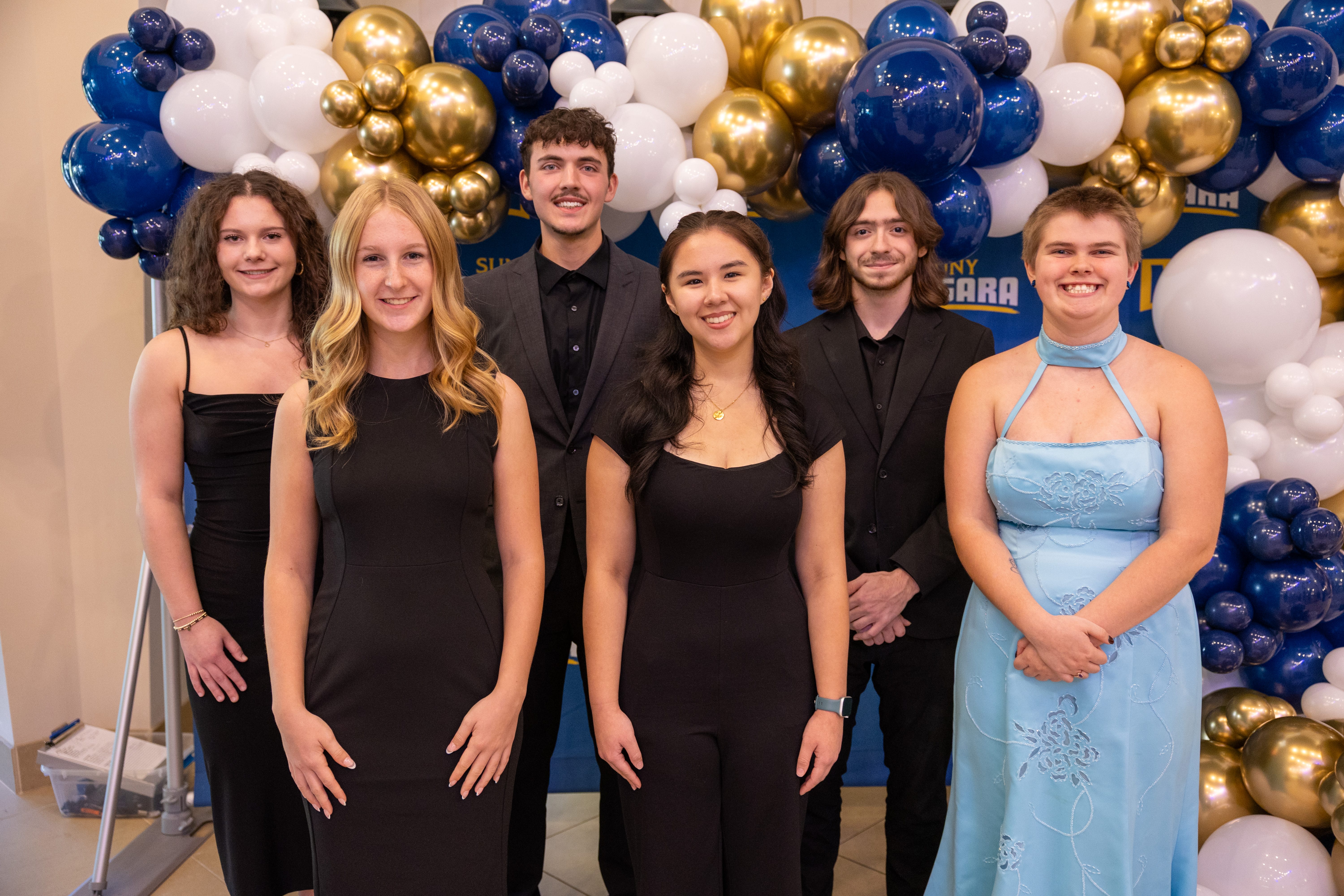 Six young adults posing and smiling in formal attire in front of a backdrop decorated with clusters of blue, gold, and white balloons, celebrating achievements like NCCC Foundation Scholarships: Spring 2025 Application Information & Deadlines.