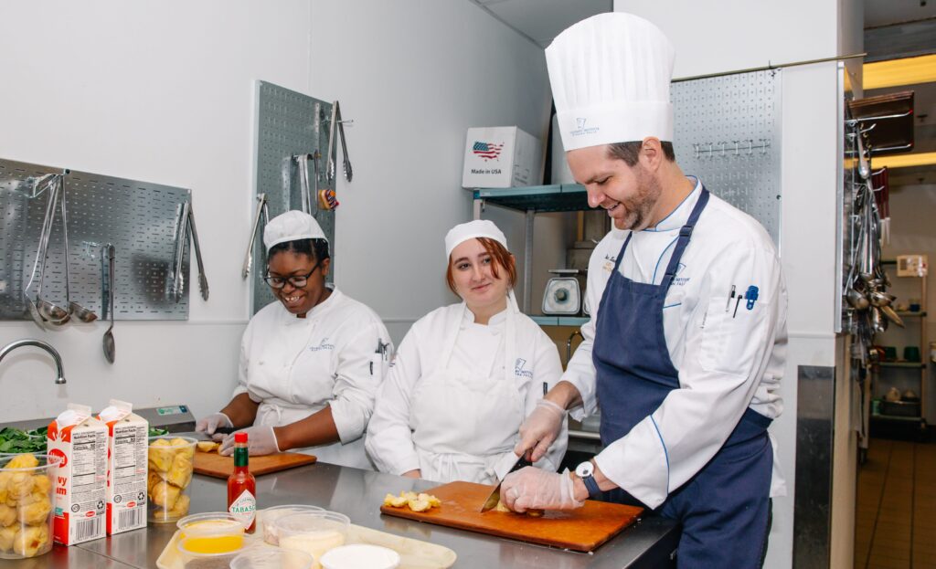 A chef and two culinary students in white uniforms and hats prepare food together in a commercial kitchen, slicing ingredients on cutting boards with various cooking supplies nearby.