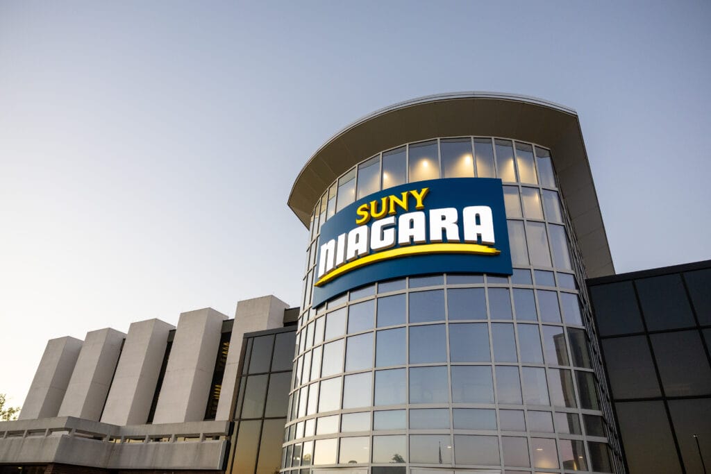 A modern glass building with a large SUNY Niagara sign on the curved facade, photographed at sunset with a clear sky in the background.