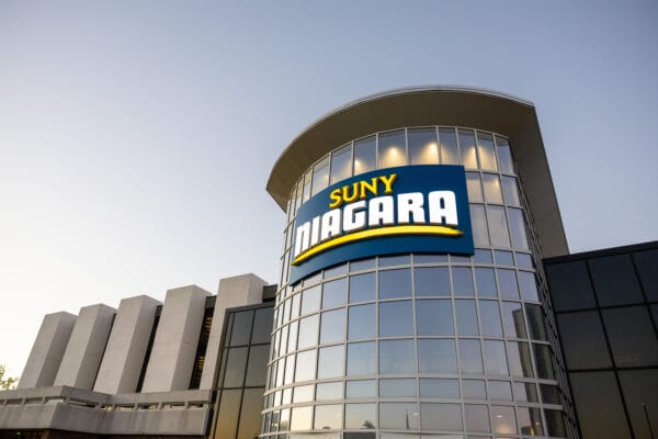 A modern glass building with a large SUNY Niagara sign on the curved facade, photographed at sunset with a clear sky in the background.
