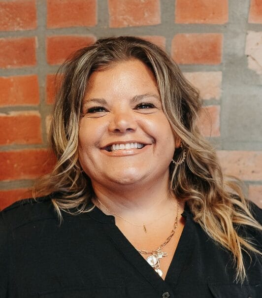 A woman with wavy, shoulder-length hair smiles in front of a brick wall. She is wearing a black shirt and layered gold necklaces.