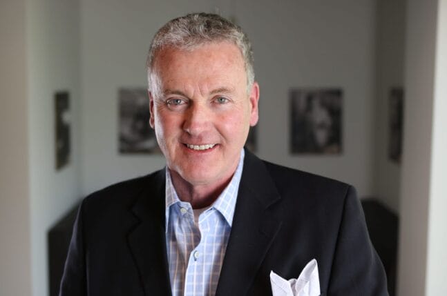 A middle-aged man with short gray hair wearing a dark suit jacket, light blue checkered shirt, and white pocket square, smiling at the camera indoors with blurred black-and-white photos on the wall behind him.