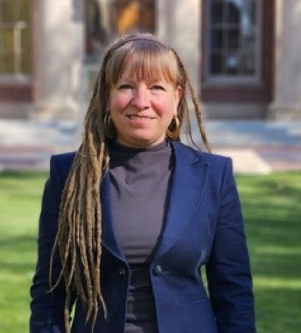 A woman with long, light brown dreadlocks and bangs, wearing a navy blazer and dark turtleneck, stands outside on a sunny day with a blurred building and green lawn in the background.