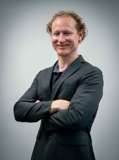 A man with curly red hair and a short beard stands against a gray background, smiling confidently with his arms crossed while wearing a black suit jacket over a dark shirt.