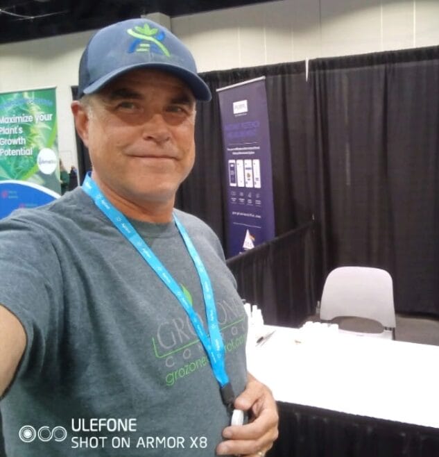 A man wearing a blue cap and gray T-shirt takes a selfie at a booth in an indoor event or expo. He is smiling, and the booth has banners and a white table with empty chairs.