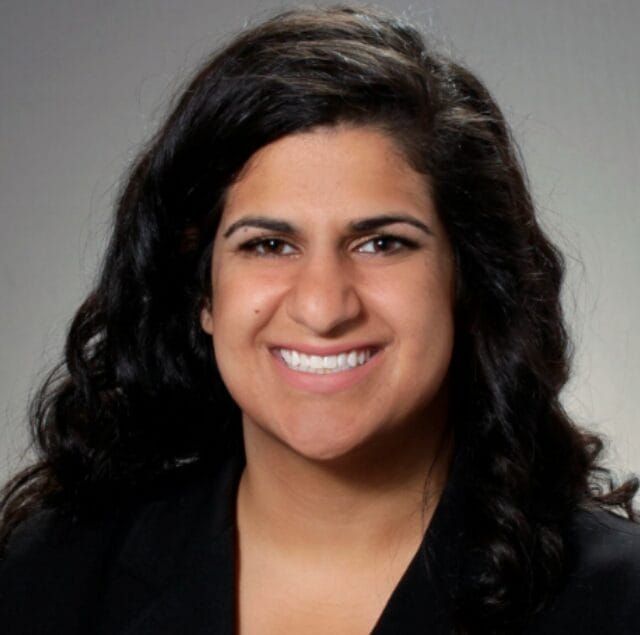 A person with long, wavy dark hair smiles at the camera. They are wearing a black blazer and are posed in front of a plain, light gray background.
