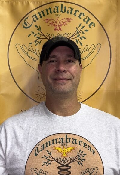 A man wearing a black cap and a white Cannabaceae T-shirt stands in front of a yellow banner with the same Cannabaceae logo, featuring hands holding branches and a phoenix.