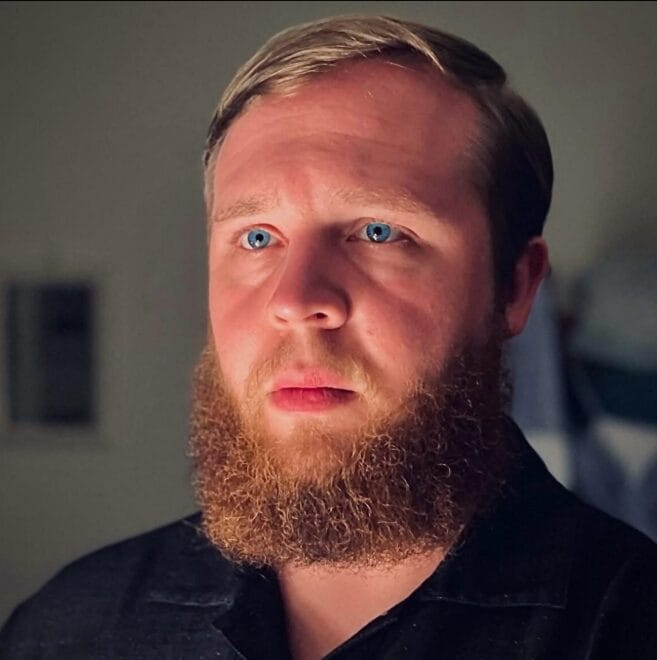 A man with a full reddish-brown beard and short light brown hair looks slightly past the camera. He is wearing a dark collared shirt and stands against a softly lit, neutral indoor background.