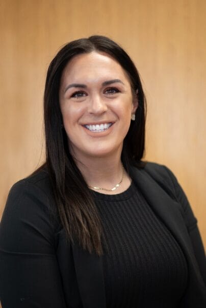 A woman with long dark hair, wearing a black blazer and black top, smiles at the camera in front of a plain light brown background.