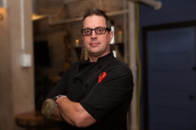 A chef with glasses and tattooed arms stands confidently with arms crossed, wearing a black chef coat in a dimly lit restaurant setting.