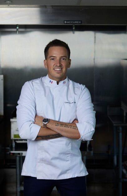 A chef in a white chef’s jacket stands with arms crossed, smiling in a commercial kitchen. He has short dark hair, a watch, and visible tattoos on his forearm. Stainless steel surfaces and kitchen equipment are in the background.