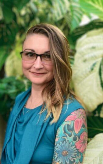 A smiling person with glasses and long, wavy, side-swept hair wears a teal top. Their right arm, featuring colorful floral tattoos, is visible. The background is filled with large, green leafy plants.