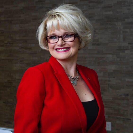 A smiling woman with short blonde hair and glasses wears a red blazer over a black top and a silver necklace, standing in front of a textured gray wall.
