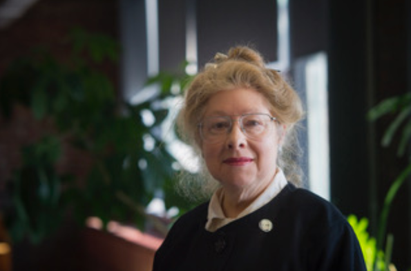 An older woman with light skin, glasses, and light-colored hair in a bun, wearing a dark jacket over a white blouse, stands indoors with plants and windows in the background.