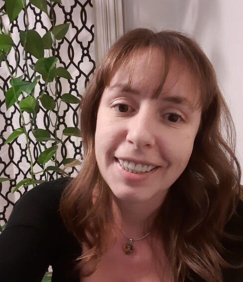 A woman with light brown hair, wearing a black top and a necklace, smiles at the camera indoors. There is a green leafy plant and a decorative black lattice-patterned screen in the background.
