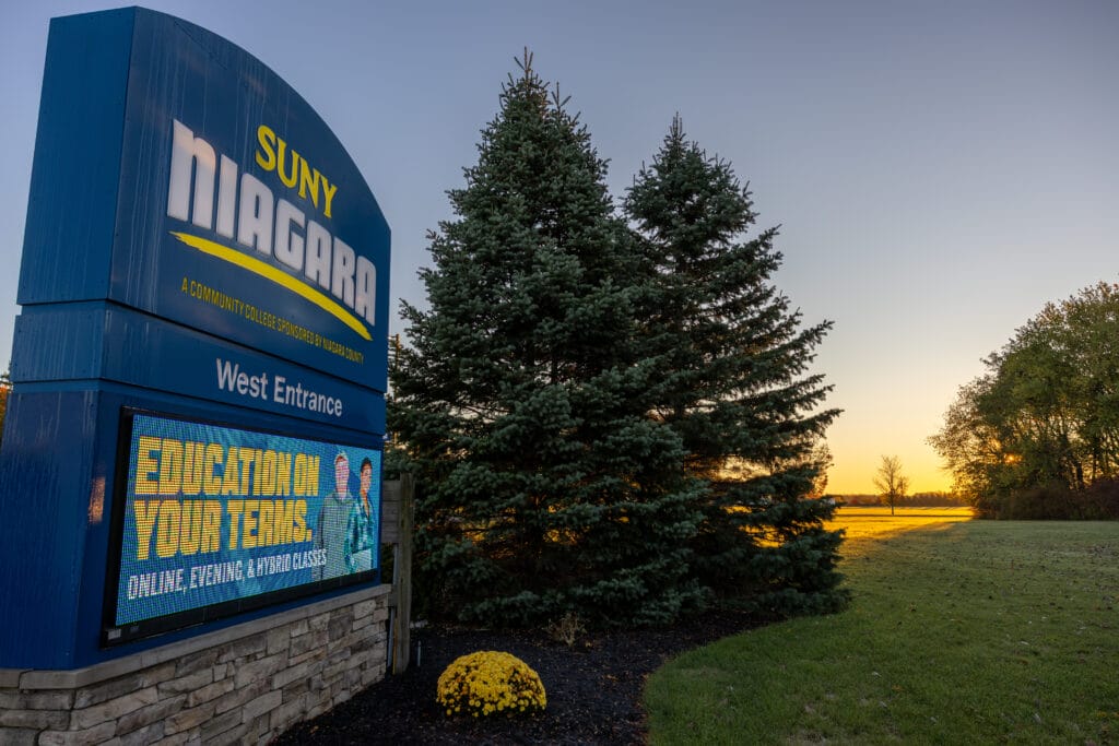 A large blue SUNY Niagara West Entrance sign stands beside two pine trees at sunset. The digital marquee reads “Education on Your Terms: Online, Evening, & Hybrid Classes.” Green grass and yellow flowers are in the foreground.