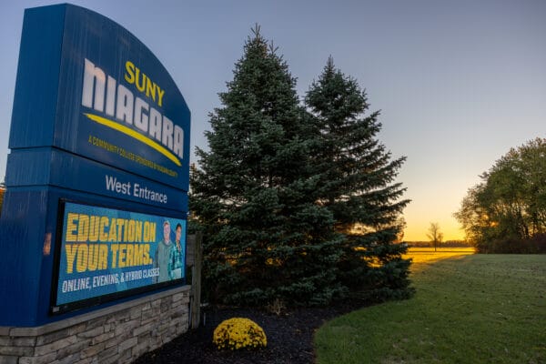 A large blue SUNY Niagara West Entrance sign stands beside two pine trees at sunset. The digital marquee reads “Education on Your Terms: Online, Evening, & Hybrid Classes.” Green grass and yellow flowers are in the foreground.