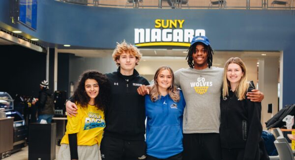 Five smiling students stand together inside a modern building with their arms around each other. Behind them is a large “SUNY Niagara” sign. They wear casual clothes, some with Niagara Wolves logos.