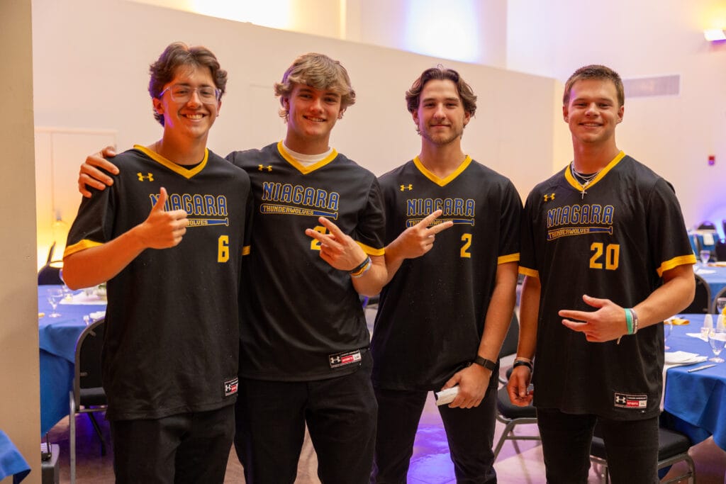 Four young men stand indoors, smiling in black Niagara Thunderwolves jerseys with yellow trim and different numbers. Three gesture with their hands. In the background, tables with blue cloths highlight their bond as Forever Connected: SUNY Niagara Alumni Association members.