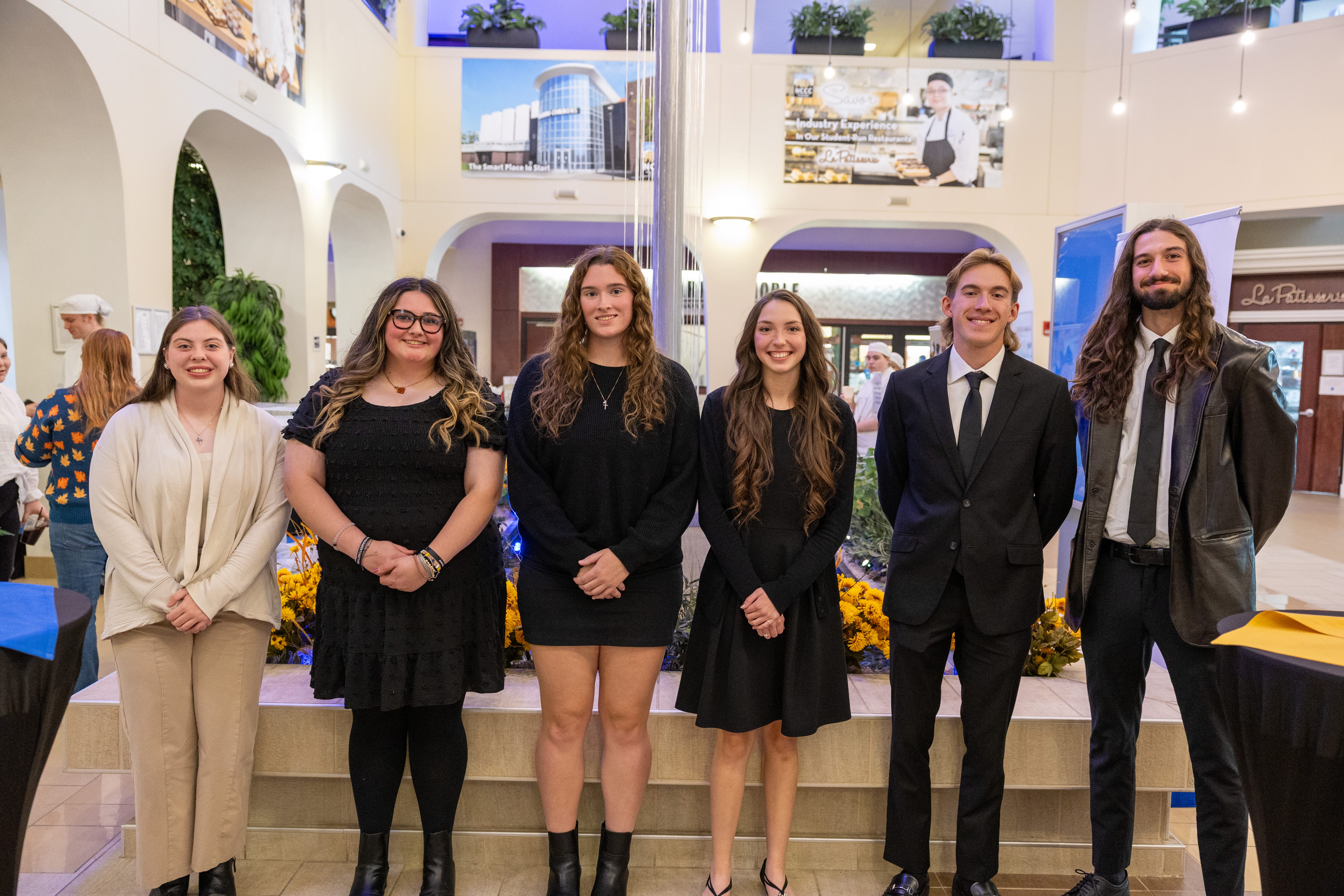 Six young adults stand side by side indoors, smiling at the camera in formal and semi-formal attire. The background features arches, plants, and a modern building display—capturing the spirit of Forever Connected: SUNY Niagara Alumni Association.