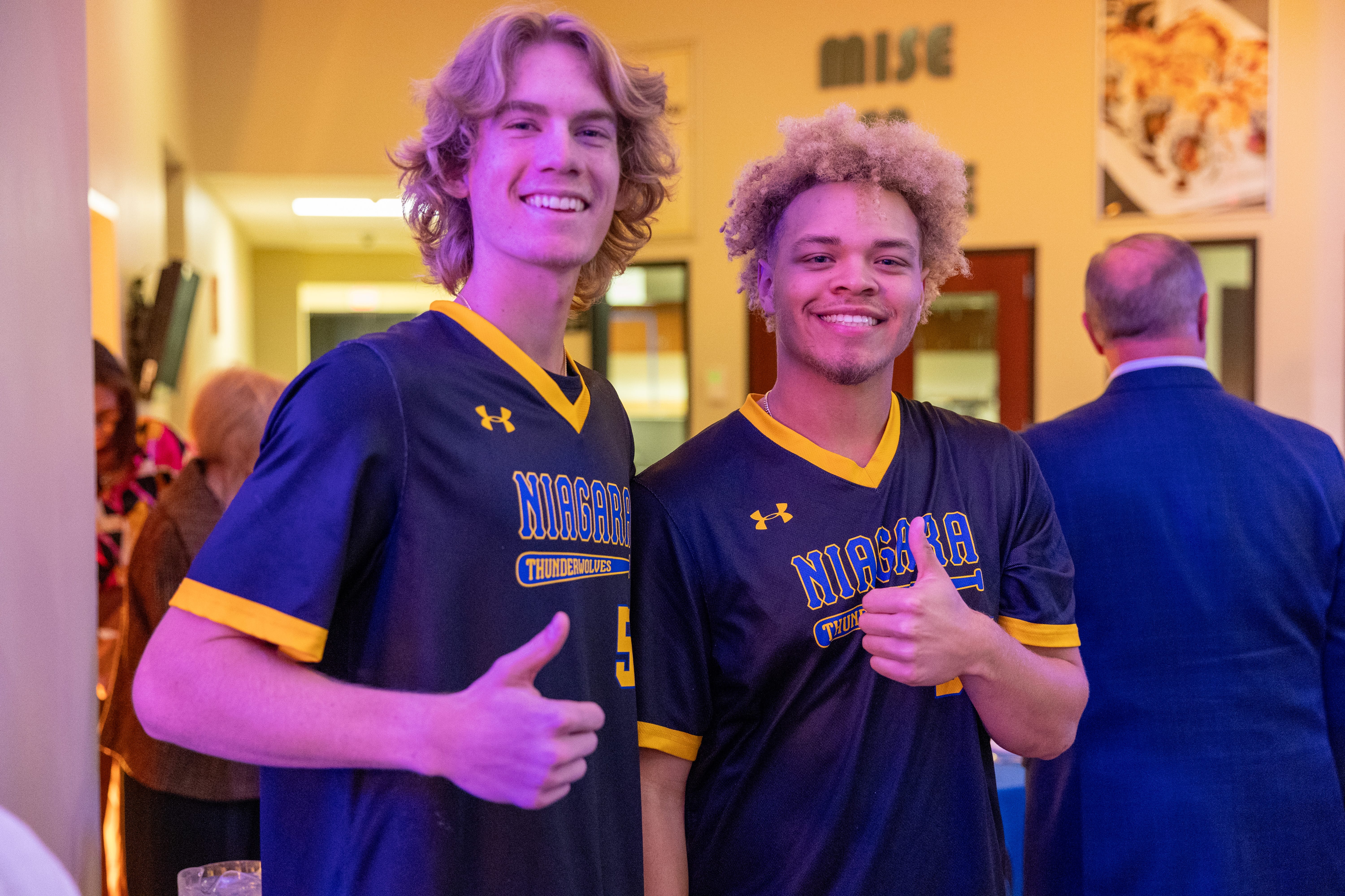 Two young men in Niagara Thunderwolves jerseys smile and give thumbs up at an indoor event, representing the Forever Connected: SUNY Niagara Alumni Association, with other people and warm lighting in the background.