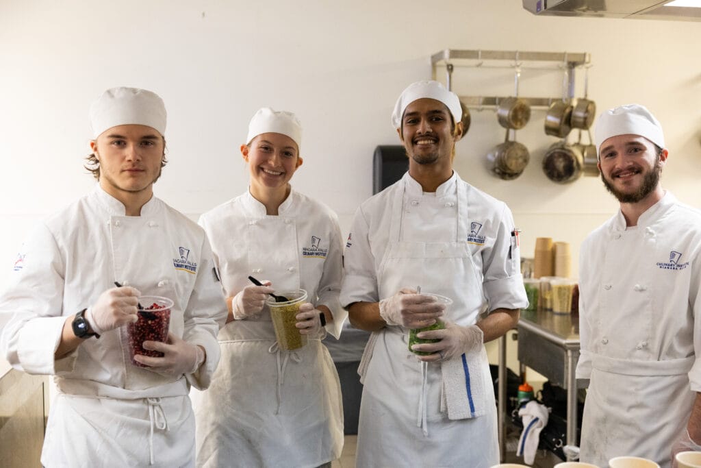 Four culinary students in white chef uniforms and hats stand in a kitchen, smiling at the camera and holding containers of chopped ingredients. Cooking utensils and metal pots are visible in the background—Forever Connected: SUNY Niagara Alumni Association.