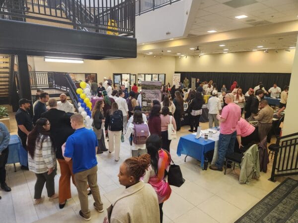 A large group of people gather at an indoor event with informational tables, banners, and balloons. Attendees are talking, collecting materials, and exploring displays in a bright, open space.