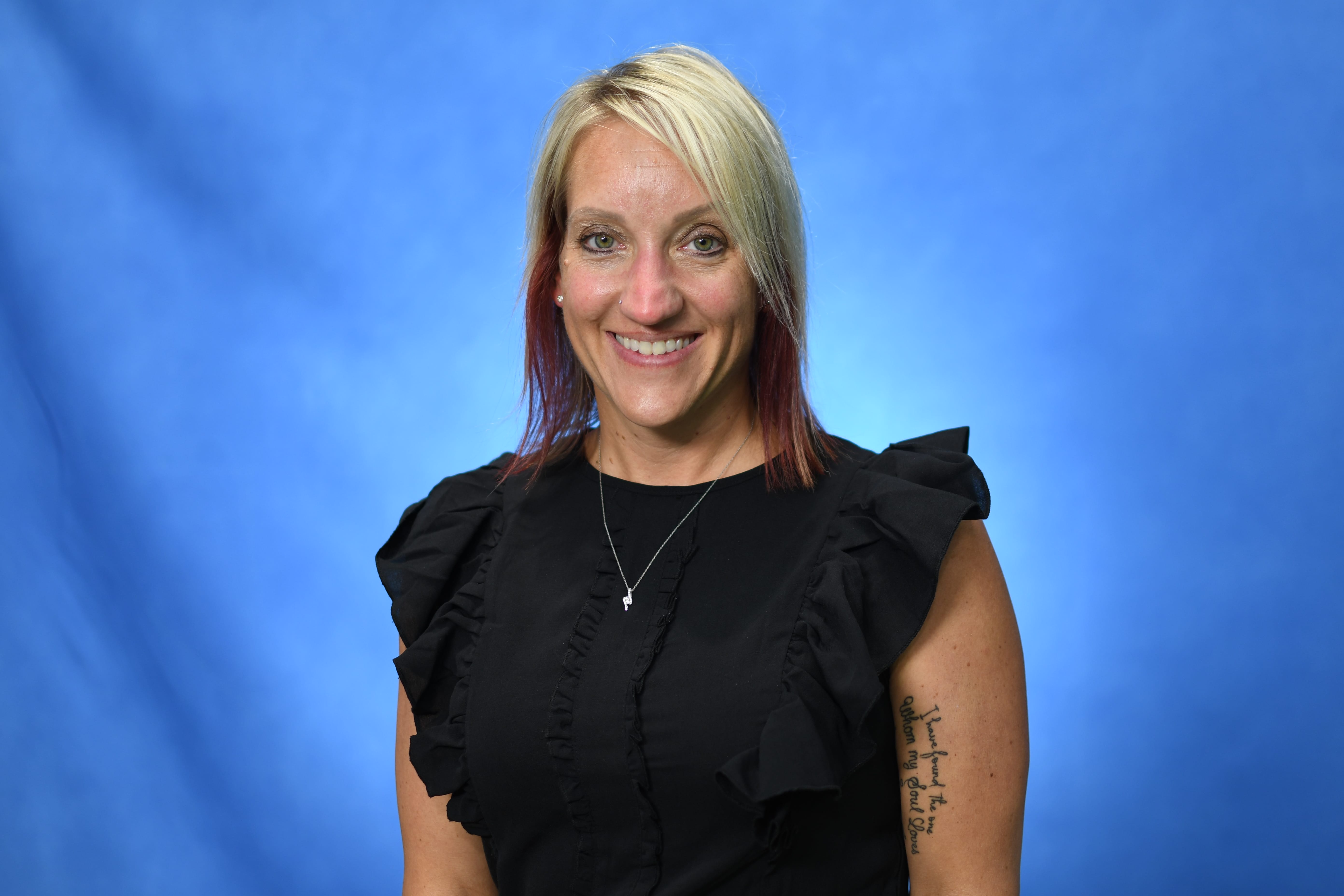 A smiling woman with shoulder-length blonde hair wearing a black ruffled sleeve top and a necklace stands in front of a solid blue background. She has a visible tattoo on her upper right arm.