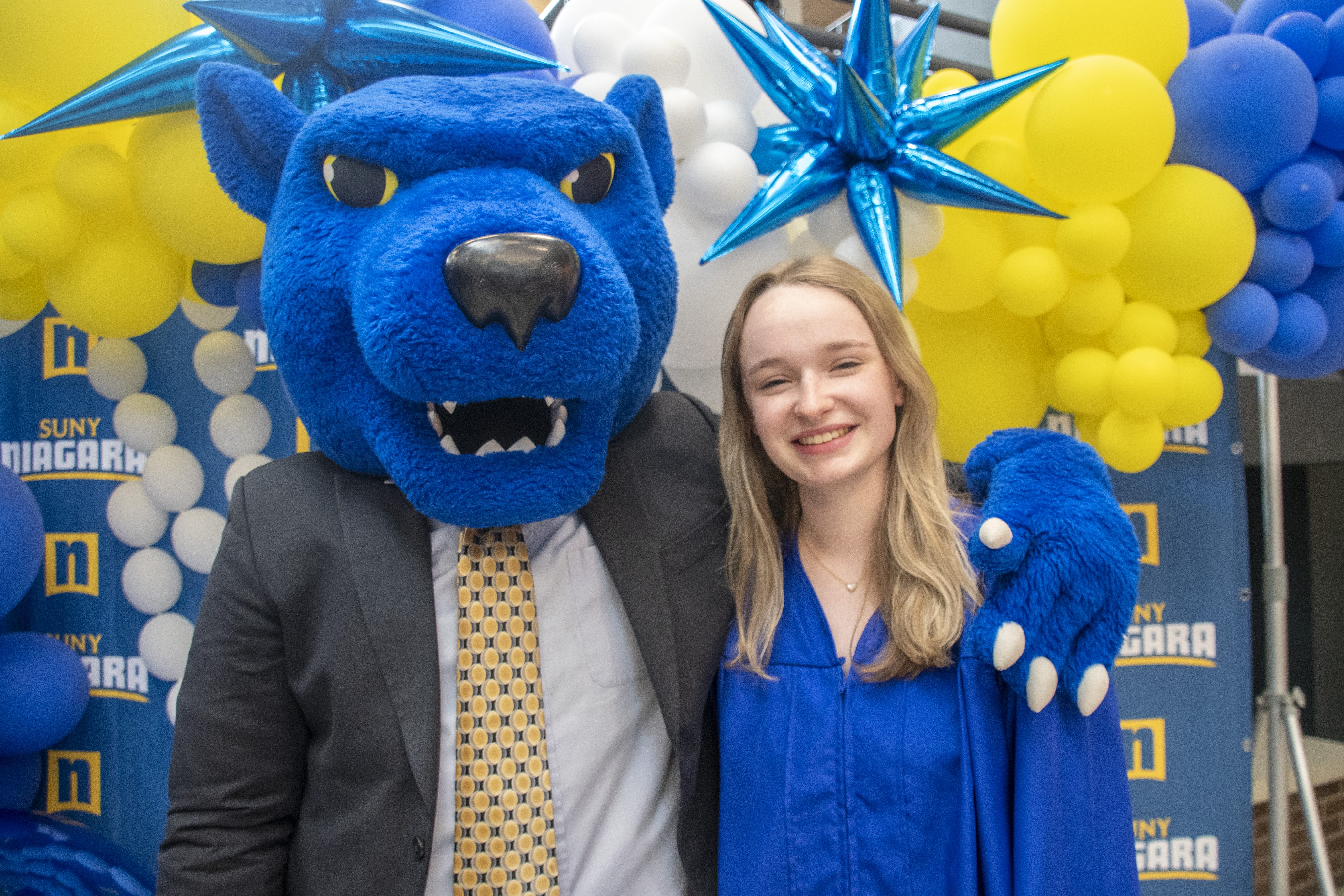 A person in a blue graduation gown stands smiling next to a blue panther mascot in a suit and tie, with colorful balloons and a SUNY Niagara backdrop—Forever Connected: SUNY Niagara Alumni Association.