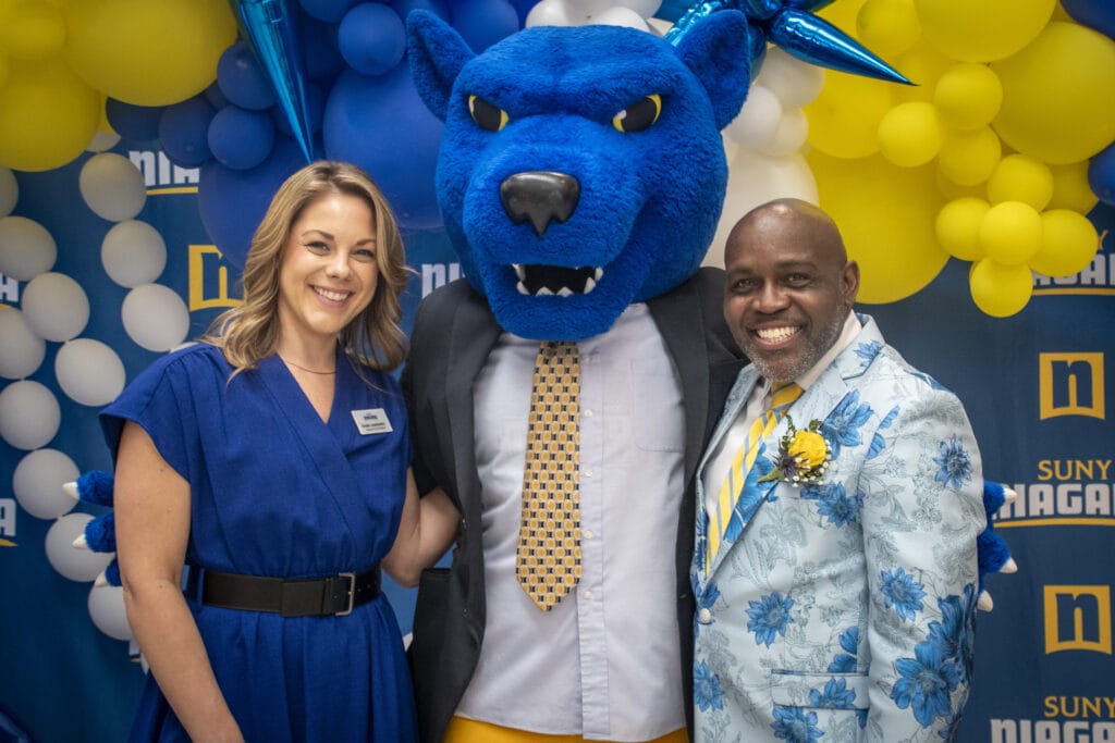 Three people pose together: a smiling woman in a blue dress, the blue wildcat mascot in a suit, and a man in a floral blue suit. Yellow and white balloons decorate the background, celebrating Forever Connected: SUNY Niagara Alumni Association.