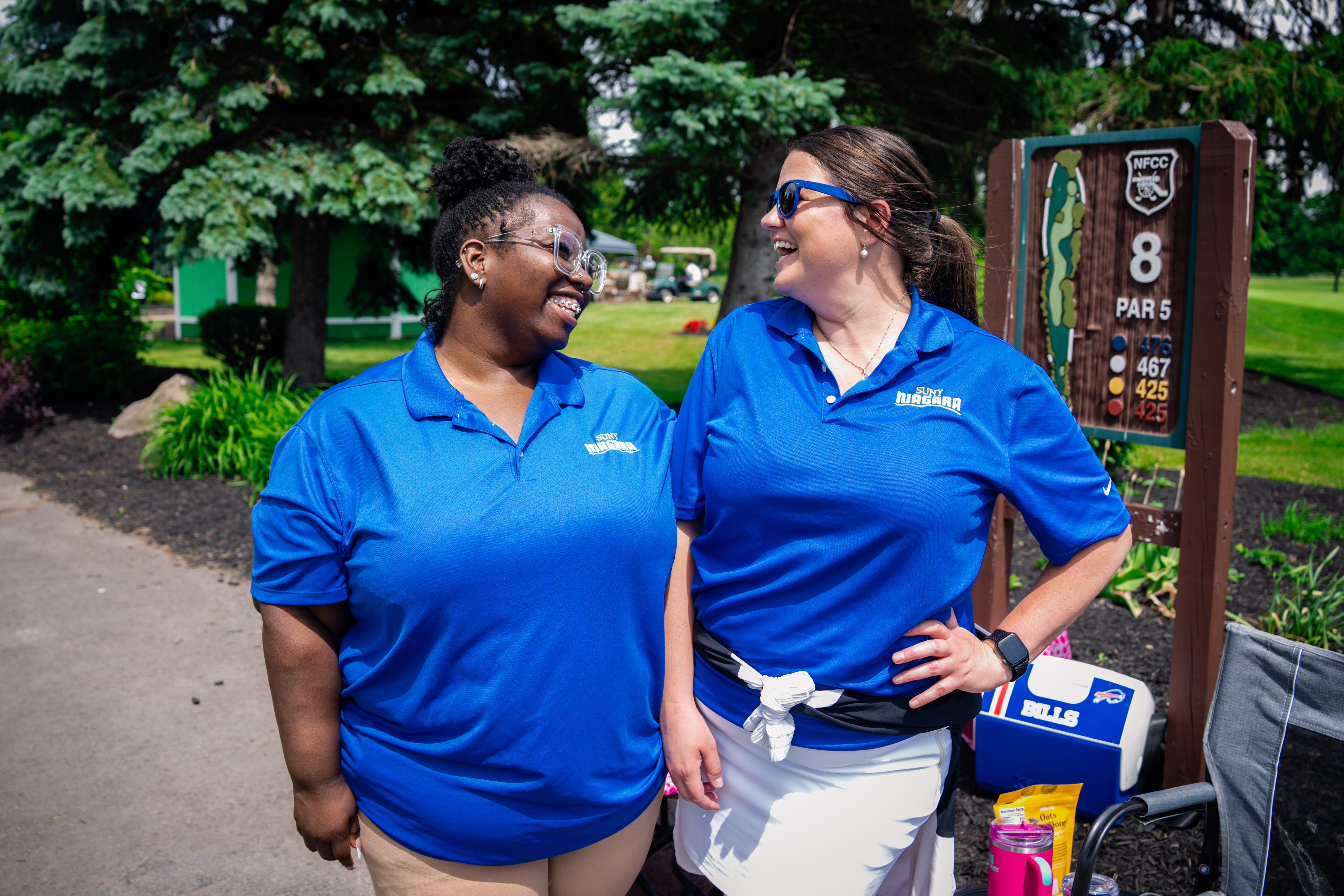 Two women in matching blue polo shirts smile at each other on a golf course near a hole 8 sign, their connection reflecting the spirit of Forever Connected: SUNY Niagara Alumni Association. Lush greenery and golf equipment complete the scene.