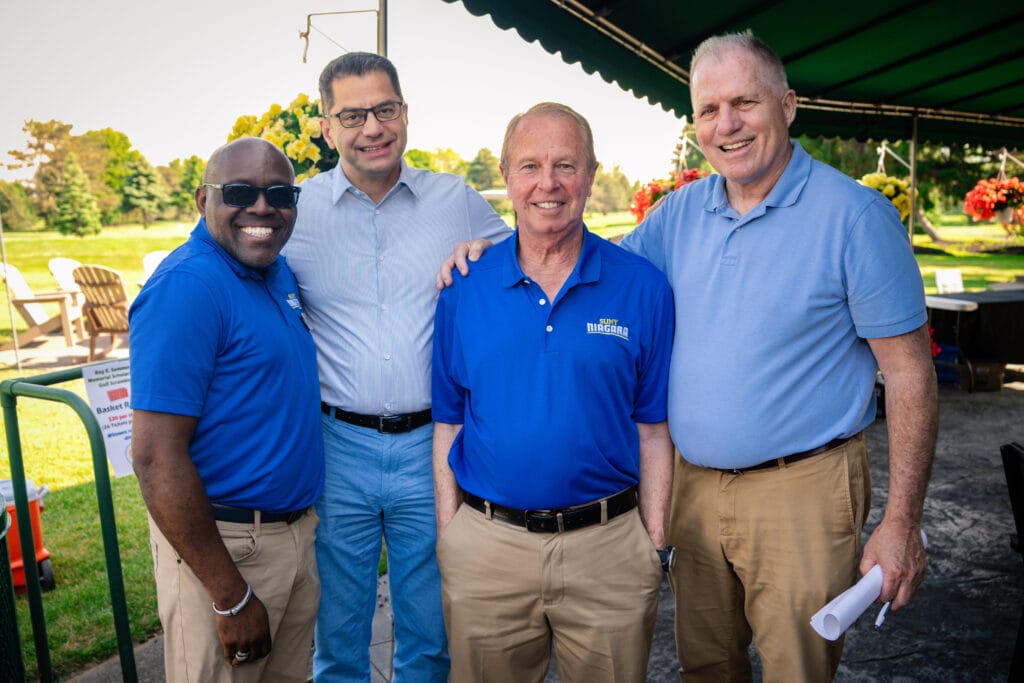 Four men are smiling and standing close together outdoors under a green canopy. Representing Forever Connected: SUNY Niagara Alumni Association, they wear coordinated blue shirts and khakis, with trees and lawn chairs in the background.