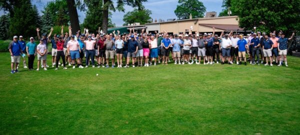 A large group of people standing together on a golf course, many with their arms raised and smiling, surrounded by green trees and a clubhouse in the background under a blue sky—Forever Connected: SUNY Niagara Alumni Association.