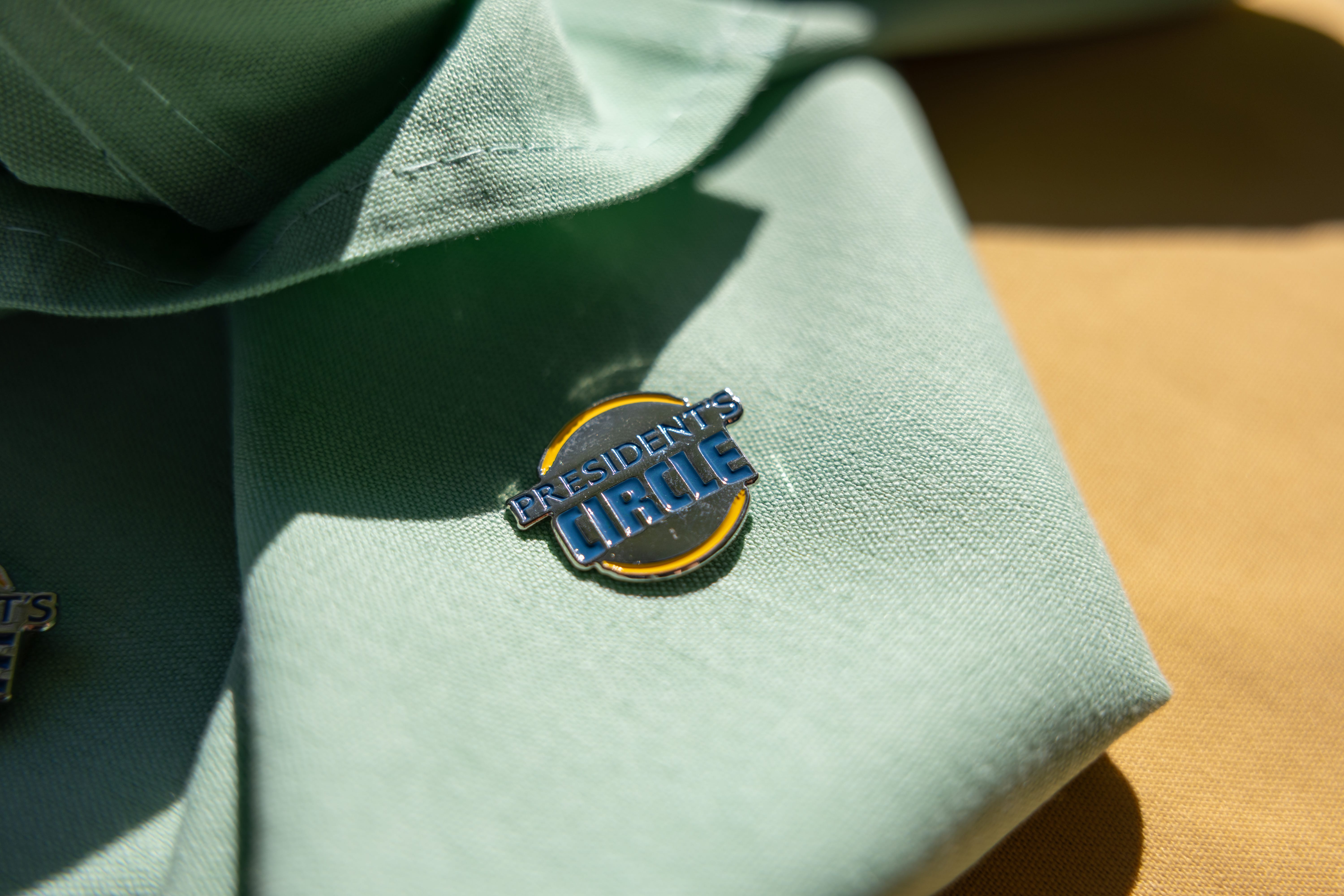 A round pin reading PRESIDENTS CIRCLE rests on a light green fabric with a folded edge and yellow background visible.
