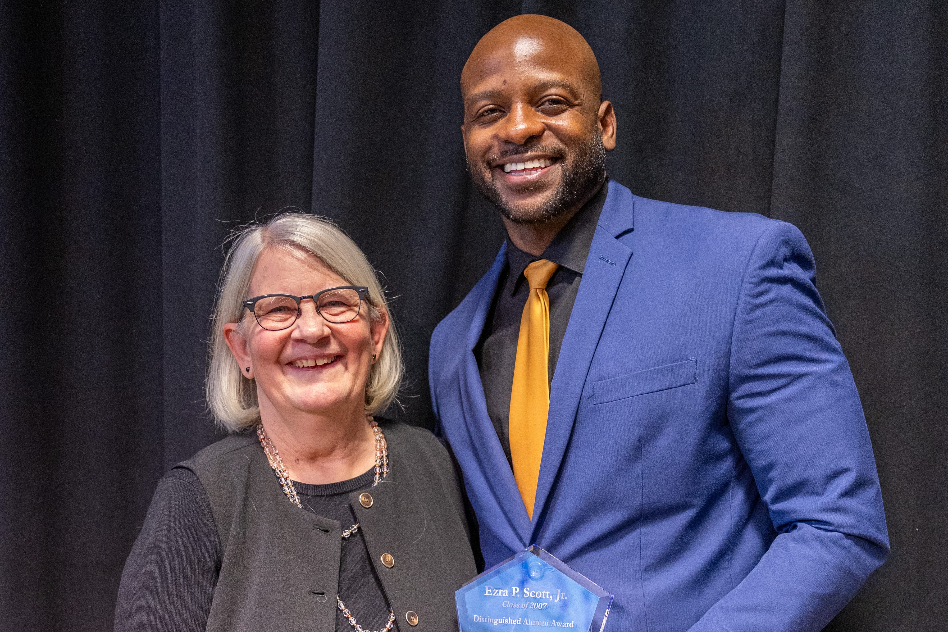 Two people stand smiling in front of a black curtain. The woman on the left has gray hair, glasses, and a gray sweater. The man on the right is bald, wearing a blue suit and gold tie, and holding a glass award.