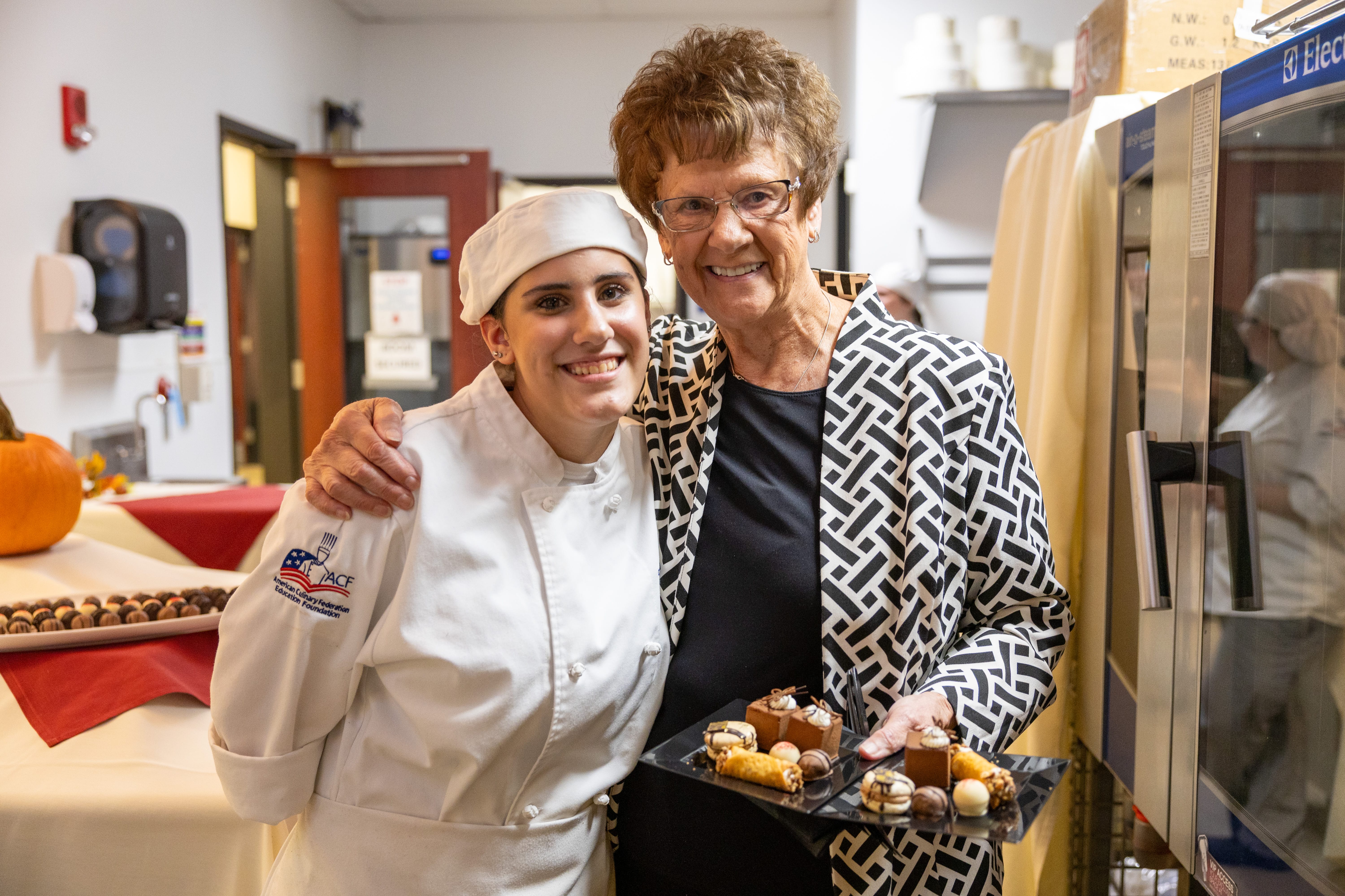 A smiling chef in a white uniform stands with an older woman holding a plate of assorted desserts. They are in a kitchen or event space with tables and food in the background.