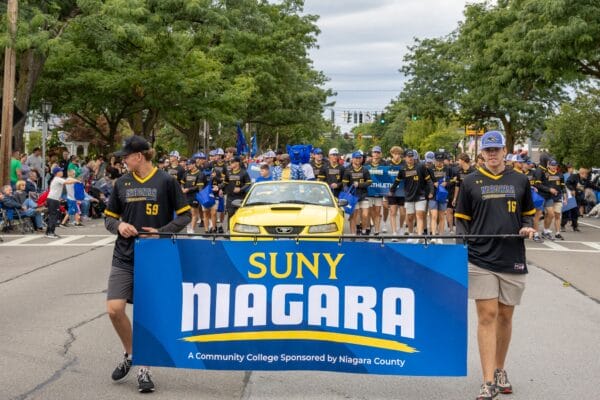 A group of people march in a parade, with two men holding a blue SUNY Niagara banner in front of a yellow car. Many others, wearing matching shirts, walk behind them. Spectators line the street under green trees.