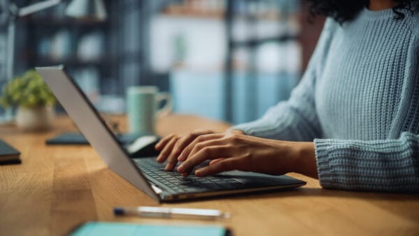 A person wearing a blue sweater types on a laptop at a wooden desk with a pen, notebook, and a mug in the background, in a cozy indoor setting.