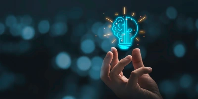 A hand reaching toward a glowing digital illustration of a lightbulb with a brain inside, symbolizing creativity and innovative ideas, against a blurred dark blue background with bokeh lights.