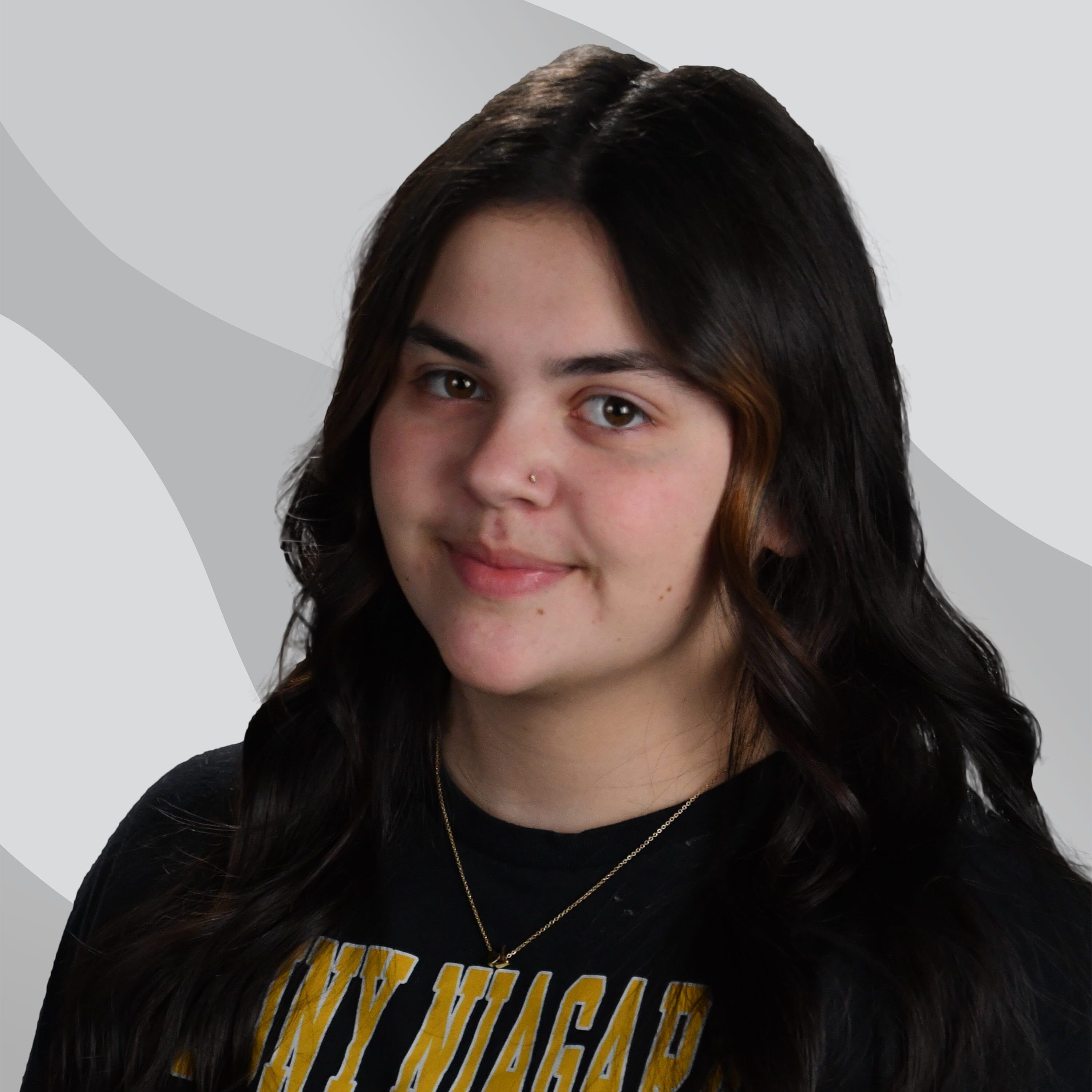 A young woman with long dark wavy hair, wearing a black SUNY Niagara Thunderwolves T-shirt, stands in front of a gray and white abstract background, smiling softly.