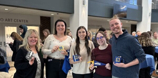 Five people stand indoors, smiling and holding mugs and prizes. They are in a bright, open area with other people and tables in the background. Signs for an art center and access center are visible on the wall.