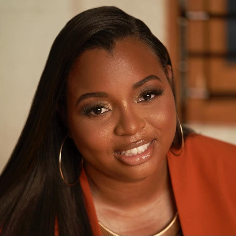 A woman with long, straight black hair and hoop earrings smiles warmly. She is wearing a rust-colored top and a gold necklace, with soft, natural makeup. The background is softly blurred.